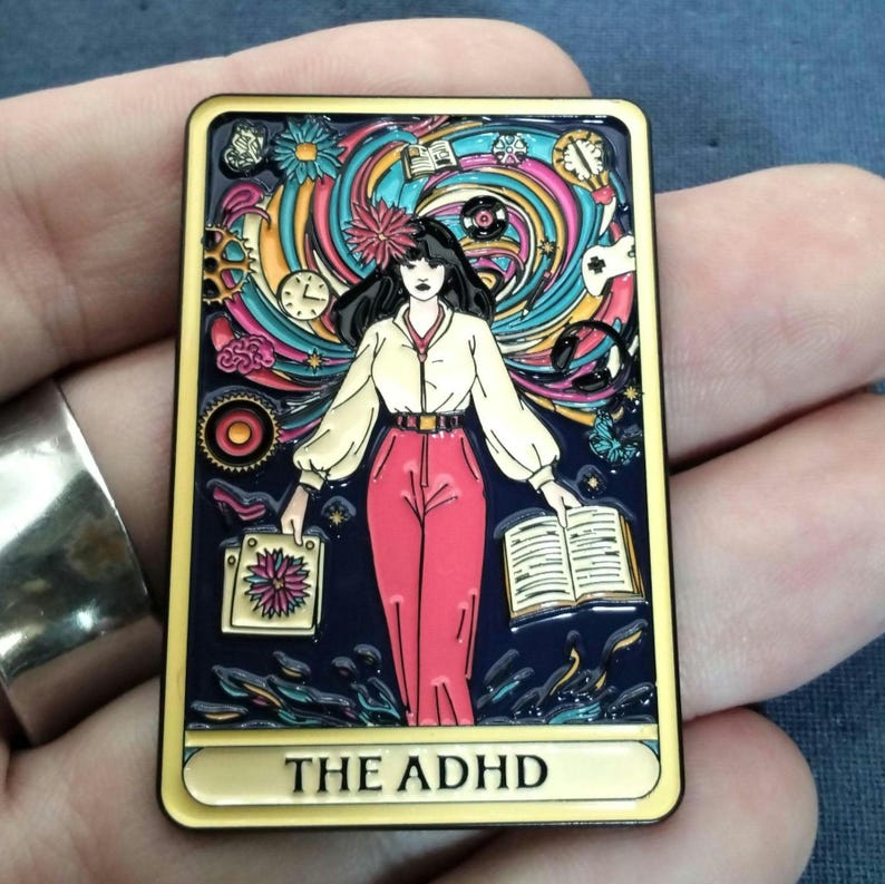 May include: A yellow-colored pin with a colorful illustration of a woman standing in a swirling vortex of colors and objects. The text "THE ADHD" is printed at the bottom of the pin like a tarot card.