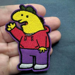 May include: An embroidered patch featuring a cartoon character with a yellow head, a green cap, a red hooded sweatshirt, and purple pants. The character is waving with one hand and has a purple border outlining the entire shape of the figure.