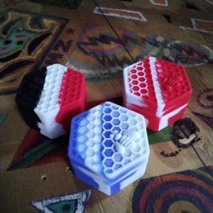 Pu&ograve; includere: Tre contenitori esagonali in silicone con un motivo a nido d'ape. I contenitori sono rosso, bianco e blu con un design di ape bianca.