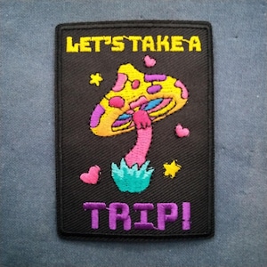 May include: Black rectangular patch with the text "LET'S TAKE A TRIP!" in yellow and purple. The patch features a colorful mushroom with yellow, pink, and purple accents, along with hearts and stars.