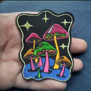 May include: Embroidered patch featuring colorful mushrooms with pink, green, and orange caps, set against a black background with yellow stars. The patch has a blue border and is likely for clothing or accessories.