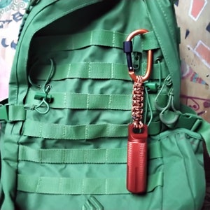 Shows A green tactical backpack with a the large orange   container attached to a orange and black carabiner and orange and black paracord lanyard.