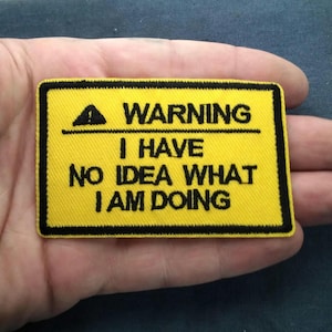 May include: Yellow rectangular patch with black border and text: "WARNING I HAVE NO IDEA WHAT I AM DOING." Features a black triangle warning symbol. The patch is held in a hand.
