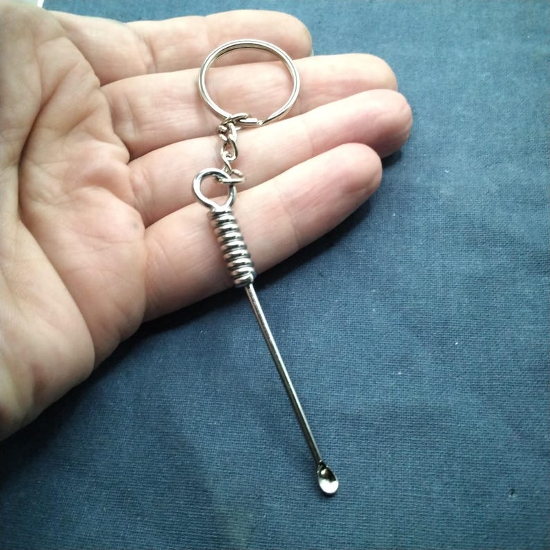 Puede incluir: Un llavero de color plateado con un peque&ntilde;o extremo en forma de cuchara. El llavero tiene un dise&ntilde;o de resorte en espiral y un anillo para sujetar llaves. El art&iacute;culo se sostiene en una mano sobre un fondo azul.