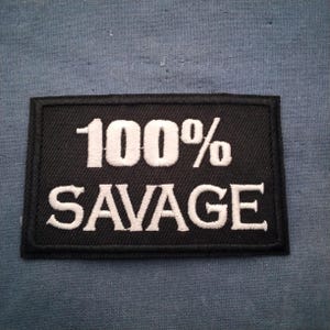 May include: A black rectangular patch with white embroidered text that reads "100% SAVAGE". The patch has a black border and is set against a blue fabric background. The text is in a bold, sans-serif font.