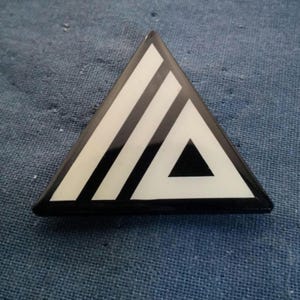 May include: A black and white triangle-shaped pin with a black triangle inside. The pin has a glossy finish.