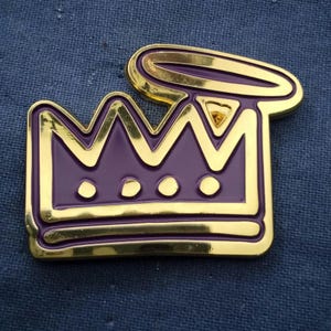 May include: A gold-colored metal pin featuring a purple crown design with a halo. The crown has three visible points and three small circles at the base. The halo is above the crown. The pin has a shiny, reflective surface.