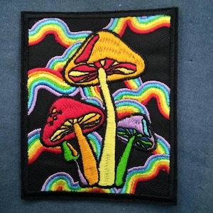 May include: Embroidered patch featuring colorful mushrooms with rainbow accents on a black background. The patch has a rectangular shape with a black border. The mushrooms are in shades of red, yellow, and purple.