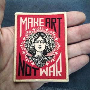May include: A red patch with the words "MAKE ART NOT WAR" in black and white. The patch features a woman's face surrounded by a floral wreath and a peace symbol. The patch has a cream-colored border.