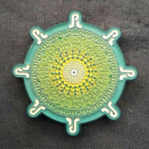 May include: A round, green decorative object with a detailed, layered design. The center features a yellow and white floral pattern, surrounded by intricate green swirls. White, stylized shapes are evenly spaced around the outer edge.