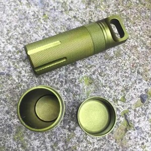 May include: A green metal pill container with a screw-on lid and a small loop on the top. The container is open and the lid is sitting next to it.