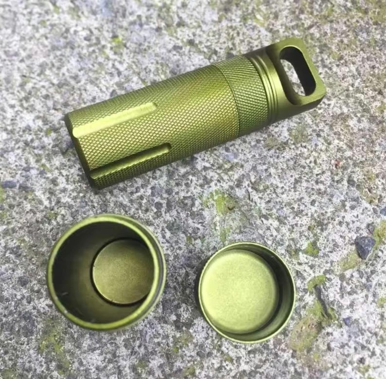 May include: A green metal pill container with a screw-on lid and a small loop on the top. The container is open and the lid is sitting next to it.