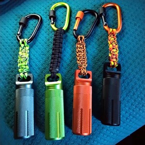 Shows Four large, cylindrical, metal pill containers with different colored tops and paracord lanyards. The containers are green, orange, black, and gray. The lanyards are green and black, orange and black, orange and yellow, and green and black.