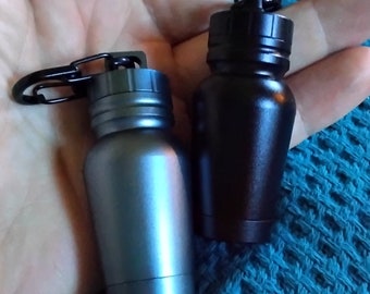 May include: Two small, cylindrical metal containers with screw-top lids and attached black carabiner clips. One container is finished in a matte metallic gray, while the other is a solid matte black. Both feature a tapered body design.
