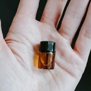 May include: A small, amber-colored glass bottle with a black cap. The bottle is held in an open hand, showcasing its size and shape. The bottle appears to contain a liquid, possibly an essential oil or perfume.