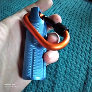 Shows the A blue metal container with an orange and black carabiner clip attached. The container has a textured surface.