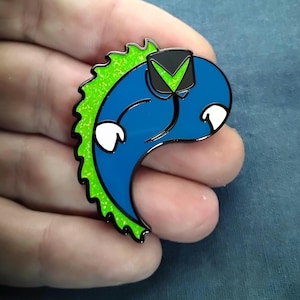 Op de afbeelding: Een blauw en groen emaille speldje met een gestileerd cartoonfiguur met een zwart-groene hoed. De pin heeft een glanzende afwerking en witte accenten. Het figuur heeft een gebogen lichaam en een groene, glitterende rug.