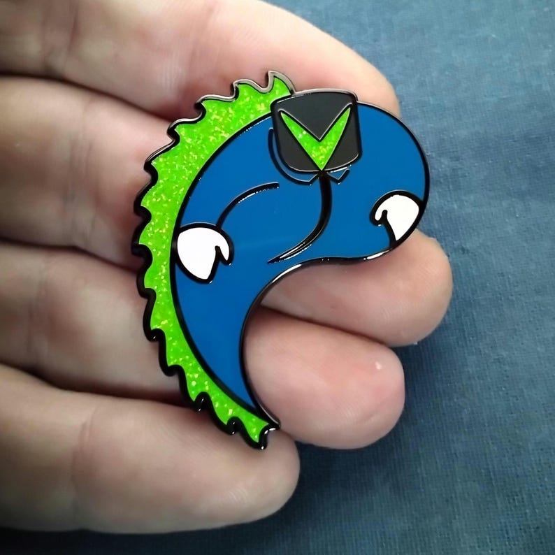 Op de afbeelding: Een blauw en groen emaille speldje met een gestileerd cartoonfiguur met een zwart-groene hoed. De pin heeft een glanzende afwerking en witte accenten. Het figuur heeft een gebogen lichaam en een groene, glitterende rug.
