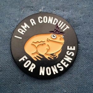 May include: A round, black button with the text "I AM A CONDUIT FOR NONSENSE" in white. The button features a cartoon frog wearing a witch hat. The frog is tan with a white belly and black outlines.