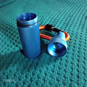 Shows a blue metal container, a cylindrical round, open-ended container. The cylindrical container has a textured surface and the lid has a textured rim.