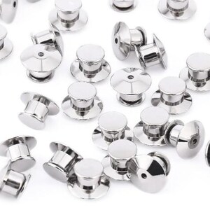 Op de afbeelding: Een verzameling glanzende, zilverkleurige pin backs, ook wel vlindersluitingen genoemd, verspreid over een wit oppervlak. Elke pin back heeft een ronde, platte bovenkant en een pin om pins te bevestigen. De backs zijn van metaal.