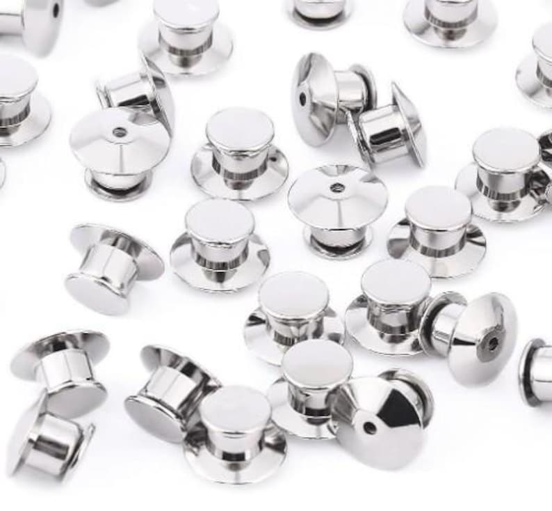 Op de afbeelding: Een verzameling glanzende, zilverkleurige pin backs, ook wel vlindersluitingen genoemd, verspreid over een wit oppervlak. Elke pin back heeft een ronde, platte bovenkant en een pin om pins te bevestigen. De backs zijn van metaal.