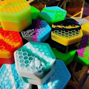 Pu&ograve; includere: Una collezione di contenitori esagonali in silicone colorati con motivi a nido d'ape e disegni di api. I contenitori sono impilati e disposti in una variet&agrave; di colori, tra cui giallo, verde, blu, rosso, nero e bianco.