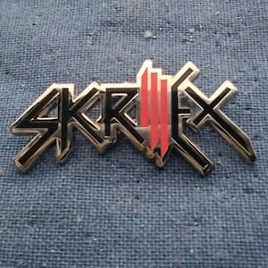May include: A silver metal pin with the word "SKRILLEX" in black letters and a red and black graphic design.