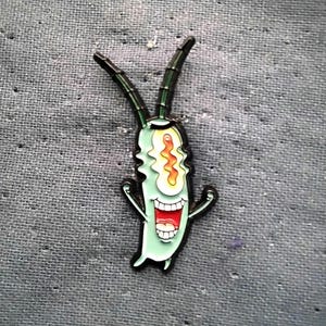 May include: A cartoon-style enamel pin featuring Plankton from Spongebob Squarepants. The pin is light green with a wide, open red mouth, white teeth, and a single orange and yellow eye. Black outlines and details complete the design.