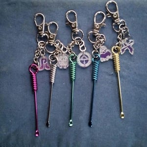 May include: A collection of keychains with metal tools and charms. The tools are in various colors, including pink, silver, green, blue, and gold. Each keychain has a clasp and a small charm with a design. The background is a solid blue color.