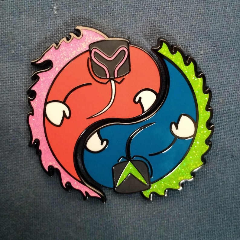 May include: A colorful enamel pin in a yin-yang design. One side is orange with pink glitter, the other is blue with green glitter. Each side has a white hands and a black and gray shape helmet.