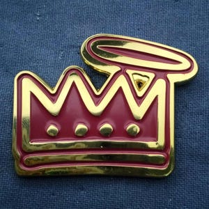 May include: A gold-colored crown pin with a burgundy center and a gold halo. The crown has a classic design with five points and three small circles at the base. The pin is likely made of metal.
