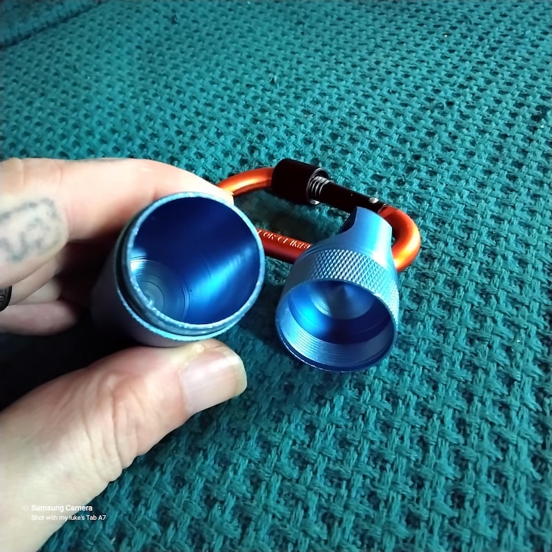 Shows a blue metal container with a carabiner clip. The container is open and show the inside of the container. The container is made of metal and have a blue finish. The carabiner clip is orange and black.