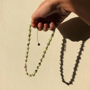 Könnte beinhalten: Eine grüne und weiße Perlenkette mit einer kleinen goldenen Kette und einem schwarzen Blumenanhänger.