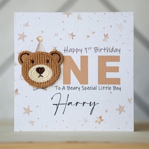 May include: A white card with a brown bear applique and the words "Happy 1st Birthday ONE To A Beary Special Little Boy Harry" in brown lettering on a white background with gold stars.