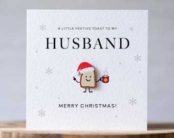 Può includere: Un biglietto d'auguri quadrato bianco con il testo A LITTLE FESTIVE TOAST TO MY HUSBAND e MERRY CHRISTMAS! e un'illustrazione centrale di una fetta di pane tostato sorridente con cappello da Babbo Natale che tiene una tazza rossa.
