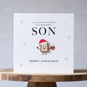 May include: A square white greeting card featuring the text A LITTLE FESTIVE TOAST TO A WONDERFUL SON and MERRY CHRISTMAS. The card displays a small illustration of a piece of toast wearing a Santa hat and holding a red mug.