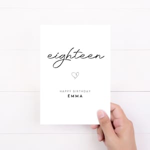 May include: A white card with black text that reads "eighteen" in cursive. Below the text is a small heart symbol and the words "Happy Birthday Emma".