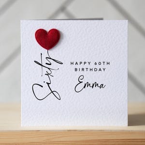 May include: A white birthday card with a red heart and the words "Sixty", "Happy 60th Birthday Emma" in black script. The card is on a wooden surface.