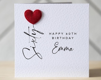 May include: A white birthday card with a red heart and the words "Sixty", "Happy 60th Birthday Emma" in black script. The card is on a wooden surface.