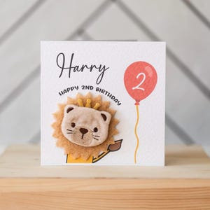 May include: A white card with the text "Harry Happy 2nd Birthday" and a red balloon with the number "2" on it. A lion with a golden crown is on the card.
