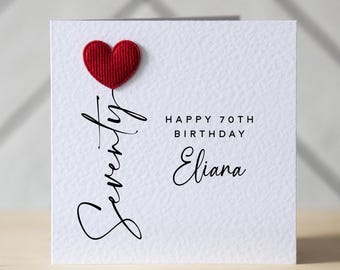 Può includere: Biglietto di auguri bianco con un cuore rosso e le scritte "Seventy", "Happy 70th Birthday" e "Eliana" in nero. Il biglietto è appoggiato su una superficie di legno.
