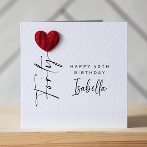 May include: A white birthday card with a red heart and the word "Forty" in cursive script. The card reads "Happy 40th Birthday Isabella" in black text. The card is on a wooden surface.