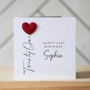 May include: A white birthday card featuring a red heart and the cursive text "Twenty One". The card also displays "Happy 21st Birthday Sophia" in black. The card is set on a wooden surface.