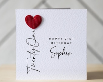 May include: A white birthday card with a red heart and the words "Twenty One" in cursive. The card also says "Happy 21st Birthday Sophia" in black text. The card is on a wooden surface.
