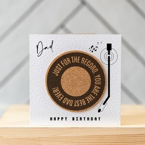 May include: A white card with the text "Dad" and "Happy Birthday" printed on it. The card features a cork record with the text "Just for the record. You are the best dad ever!" printed on it. A black and white illustration of a record player arm is shown next to the record.