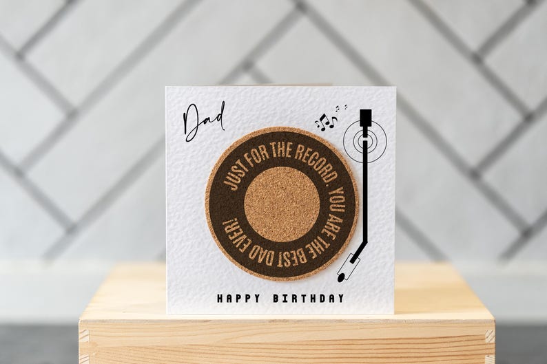 May include: A white card with the text "Dad" and "Happy Birthday" printed on it. The card features a cork record with the text "Just for the record. You are the best dad ever!" printed on it. A black and white illustration of a record player arm is shown next to the record.