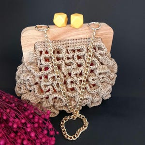 May include: A raffia clutch bag with a wooden frame and gold chain strap. The bag has a woven design and a heart-shaped pendant. Two yellow geometric shapes sit on the wooden frame. The background is black.