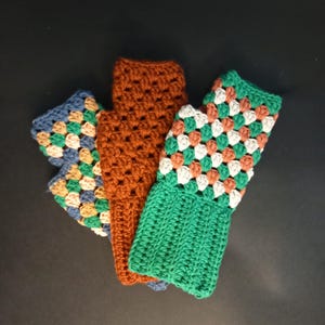 以下が含まれることがあります： 異なる色と模様のクロシェ編み食器拭きが3枚。1枚は茶色、1枚は白とオレンジのアクセントが付いた緑色、もう1枚は白とオレンジのアクセントが付いた青色です。