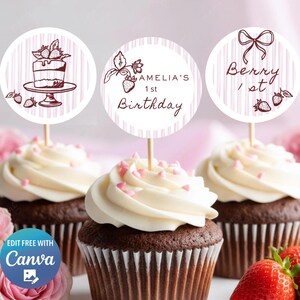 Può includere: Tre cupcake al cioccolato con glassa bianca e zuccherini rosa. Ogni cupcake ha un topper rotondo a tema fragola. Uno dice "Amelia's 1st Birthday", un altro dice "Berry 1st". Fragole fresche in primo piano.
